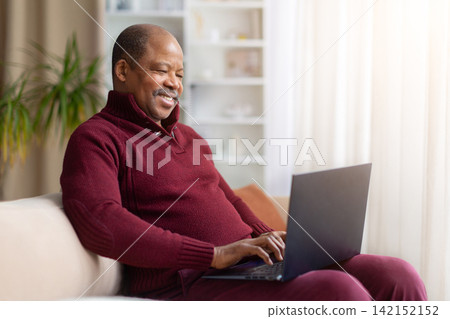 Smiling mature Black man sitting on sofa at home and using laptop 142152152
