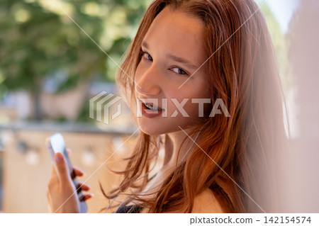 Red haired Gen Z teenager with braces gives confident playful side glance while holding smartphone. Concept of authentic teen confidence and cheeky self assured attitude in everyday lifestyle moment. Red haired Gen Z teenager with braces gives confident playful side glance while holding smartphone. Concept of authentic teen confidence and cheeky self assured attitude in everyday lifestyle moment. 142154574