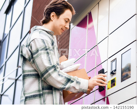 Man using smartphone at automated parcel locker for contactless delivery pickup. Modern self-service terminal for online shopping logistics and courier service. Man using smartphone at automated parcel locker for contactless delivery pickup. Modern self-service terminal for online shopping logistics and courier service. 142155190