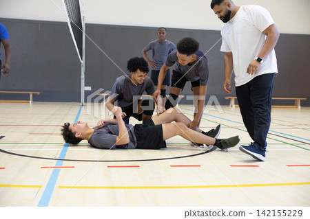 Teenage male players with coach kneeling around injured teammate lying on volleyball court near net Teenage male players with coach kneeling around injured teammate lying on volleyball court near net 142155229