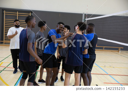 African American male teammates in jerseys huddling and smiling on court with net, coach clapping African American male teammates in jerseys huddling and smiling on court with net, coach clapping 142155233