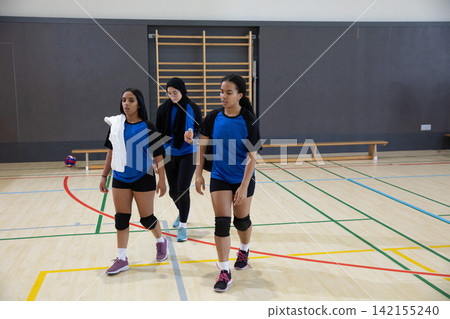 Three female volleyball teammates walking across school gym court in jerseys, volleyball nearby Three female volleyball teammates walking across school gym court in jerseys, volleyball nearby 142155240