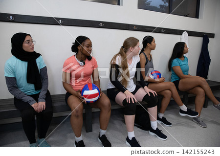 Female volleyball team sitting on bench in locker room wearing sportswear holding volleyballs Female volleyball team sitting on bench in locker room wearing sportswear holding volleyballs 142155243