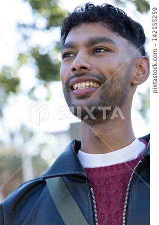 Indian man smiling, wearing black leather jacket and maroon sweater with green strap in park Indian man smiling, wearing black leather jacket and maroon sweater with green strap in park 142155259