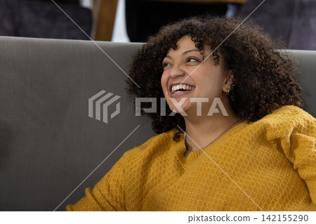 Adult African American woman laughing, sitting on sofa in mustard knit, hoop earrings, copy space 142155290