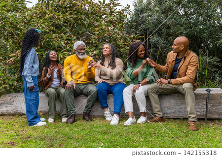 African American family sitting on wooden log bench in park passing small brown seeds in sweaters 142155318