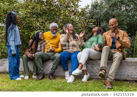 African American family in knit sweaters laughing, sitting on wooden log bench on lawn holding cup 142155319