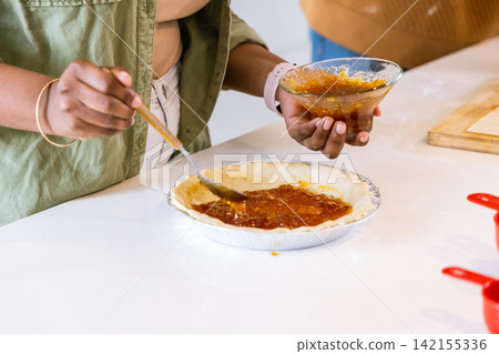 African American woman in green jacket and companion spreading jam into aluminum pie tin on counter African American woman in green jacket and companion spreading jam into aluminum pie tin on counter 142155336
