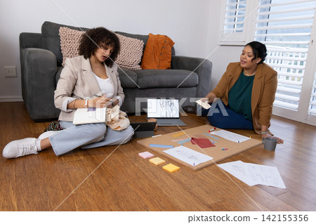 Female colleagues in blazers sitting on wood floor in living room, working over laptop and sketches Female colleagues in blazers sitting on wood floor in living room, working over laptop and sketches 142155356