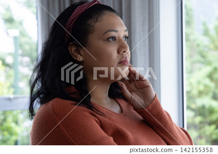 Asian woman in mid-30s sitting by large window at home, wearing rust sweater, red headband Asian woman in mid-30s sitting by large window at home, wearing rust sweater, red headband 142155358