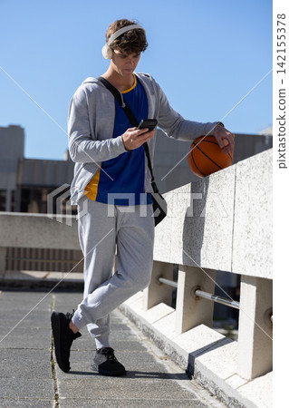 Late teen male leaning on rooftop parapet, checking phone and holding basketball, using headphones Late teen male leaning on rooftop parapet, checking phone and holding basketball, using headphones 142155378