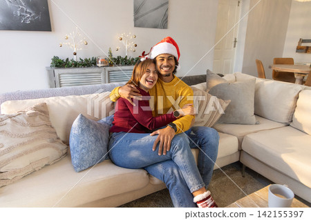 Couple sitting on sectional sofa, man wearing Santa hat holding partner, holiday decorations behind 142155397