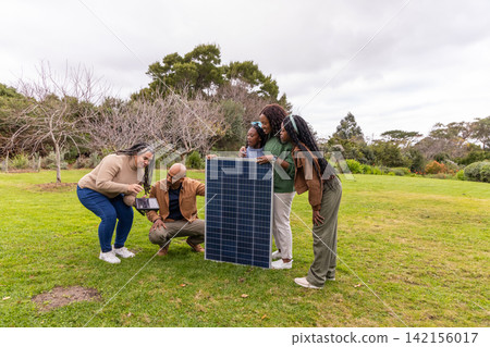 African American family kneeling and examining tablet by vertical solar panel on grassy lawn 142156017