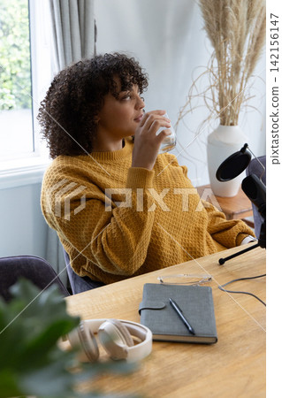 African American female sitting in home studio wearing mustard sweater holding glass by microphone African American female sitting in home studio wearing mustard sweater holding glass by microphone 142156147
