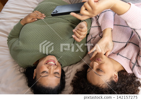 African American female friends lying on bed in knit sweaters posing for selfie, holding smartphone African American female friends lying on bed in knit sweaters posing for selfie, holding smartphone 142157275