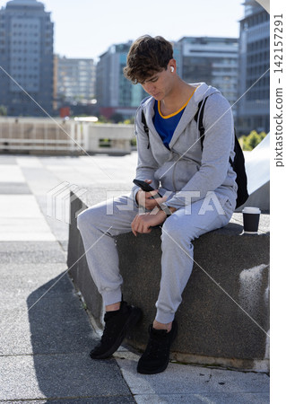 Teenage male sitting on ledge at plaza checking smartphone while wearing grey tracksuit and earbuds Teenage male sitting on ledge at plaza checking smartphone while wearing grey tracksuit and earbuds 142157291