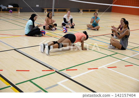 Female volleyball team watching teammate doing push-ups on gym court wearing coral top, volleyballs Female volleyball team watching teammate doing push-ups on gym court wearing coral top, volleyballs 142157404