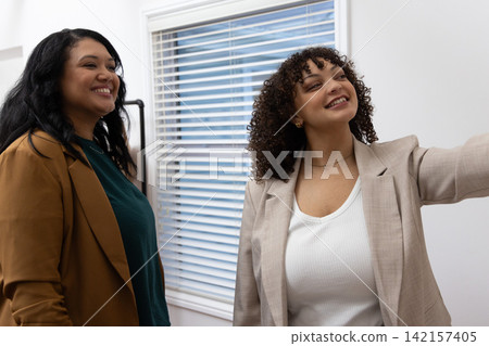 Diverse female colleagues smiling and standing close together in office with blazers, blinds, rack Diverse female colleagues smiling and standing close together in office with blazers, blinds, rack 142157405