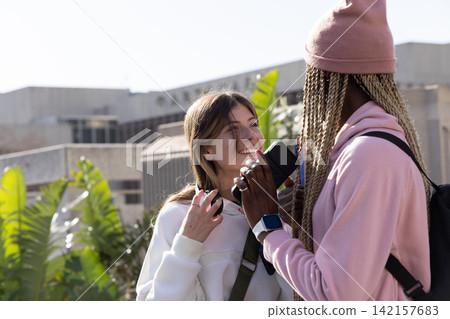 Diverse female friends standing on campus, holding smartphone with headphones and backpack Diverse female friends standing on campus, holding smartphone with headphones and backpack 142157683
