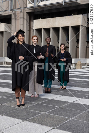 Diverse female graduates posing on tiled campus plaza wearing black gowns and adjusting mortarboard 142157689