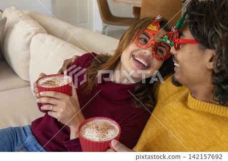 Couple woman leaning on Indian man, wearing holiday glasses, holding red frothy mugs, showing ring Couple woman leaning on Indian man, wearing holiday glasses, holding red frothy mugs, showing ring 142157692