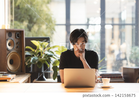 一位二十多歲的男子坐在燈光明亮的房間裡的桌子前,打開筆記型電腦,陷入沉思。 一位二十多歲的男子坐在燈光明亮的房間裡的桌子前,打開筆記型電腦,陷入沉思。 142158675