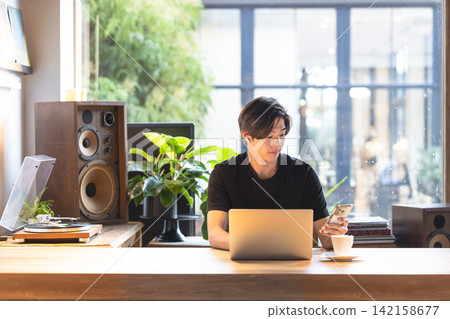 A man in his 20s using his smartphone while working on his computer. 142158677