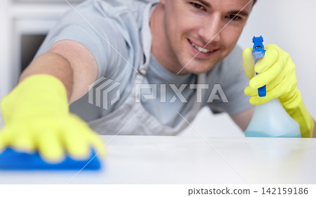 Spray, kitchen counter and and man with smile, cleaning and housekeeping in home or hotel. Housework, housekeeper or male cleaner service wiping dirt on table with cloth, soap and bottle in apartment Spray, kitchen counter and and man with smile, cleaning and housekeeping in home or hotel. Housework, housekeeper or male cleaner service wiping dirt on table with cloth, soap and bottle in apartment 142159186
