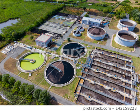 Wastewater treatment facility shows large tanks and buildings surrounded by green areas and fields during daylight hours Wastewater treatment facility shows large tanks and buildings surrounded by green areas and fields during daylight hours 142159228