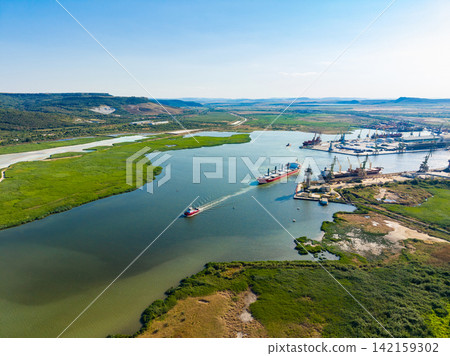 Ships navigate a river near a port with green banks and mountains in the background during a clear day Ships navigate a river near a port with green banks and mountains in the background during a clear day 142159302