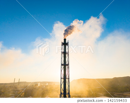 Factory smokestack releases smoke into the sky at sunset with industrial structures in the background Factory smokestack releases smoke into the sky at sunset with industrial structures in the background 142159433