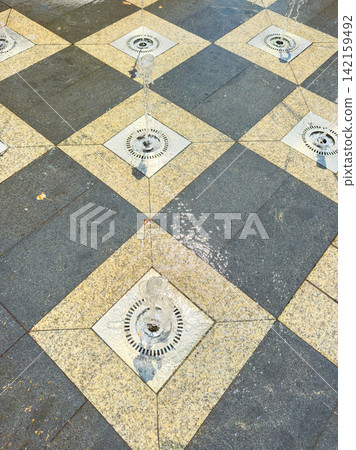 Water jets spray in a pattern on a tiled area during a sunny day in a public space Water jets spray in a pattern on a tiled area during a sunny day in a public space 142159492
