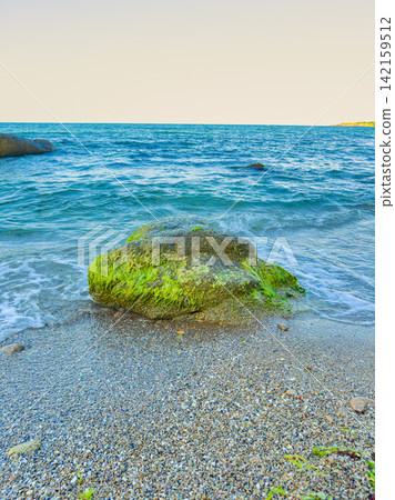 Waves crash gently on a rock covered with green moss by the shore at a beach during the day Waves crash gently on a rock covered with green moss by the shore at a beach during the day 142159512