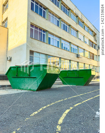 Large green waste containers sit on the asphalt surface near a building during daytime in an urban area Large green waste containers sit on the asphalt surface near a building during daytime in an urban area 142159684