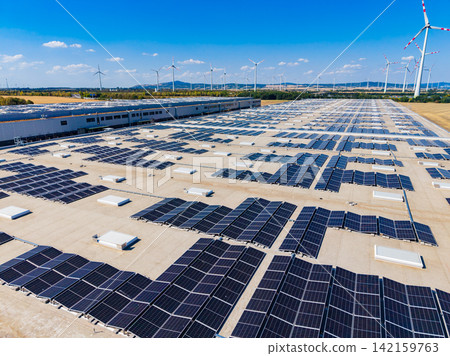 Solar panels cover the roof of a large building in a sunny location with wind turbines in the background Solar panels cover the roof of a large building in a sunny location with wind turbines in the background 142159763