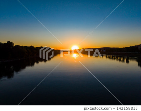 Sunset over river, showing still water reflecting orange and blue sky, with silhouette of trees along the shore during evening Sunset over river, showing still water reflecting orange and blue sky, with silhouette of trees along the shore during evening 142159813