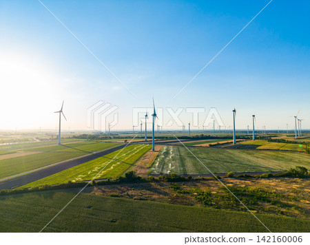 Wind turbines stand tall in the green fields on a sunny day in the countryside while the sun rises in the background Wind turbines stand tall in the green fields on a sunny day in the countryside while the sun rises in the background 142160006