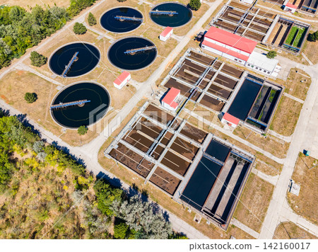 View of a wastewater treatment facility showing several large circular and rectangular tanks surrounded by green space and buildings in a city View of a wastewater treatment facility showing several large circular and rectangular tanks surrounded by green space and buildings in a city 142160017