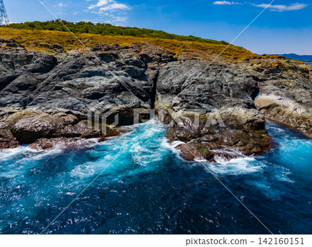 Rocks along the shore with blue water and green hills in the background on a sunny day 142160151