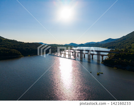 Bridge spans over lake with surrounding mountains and sun shining in the clear sky during daytime Bridge spans over lake with surrounding mountains and sun shining in the clear sky during daytime 142160233