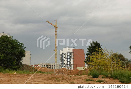 Active urban development site under a cloudy sky with red brown dirt foreground, chain link fence, and dirt piles. Active urban development site under a cloudy sky with red brown dirt foreground, chain link fence, and dirt piles. 142160414