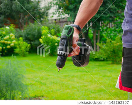 Close-up of a hand wearing a black watch gripping a green cordless drill against a blurred green lawn and garden background. Close-up of a hand wearing a black watch gripping a green cordless drill against a blurred green lawn and garden background. 142162507