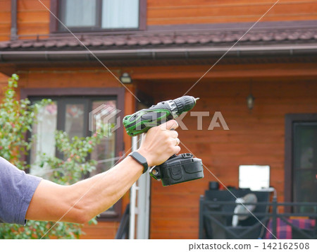 Arm and hand wearing a smartwatch holding a green cordless drill in front of a wooden cabin wall with a window. Arm and hand wearing a smartwatch holding a green cordless drill in front of a wooden cabin wall with a window. 142162508