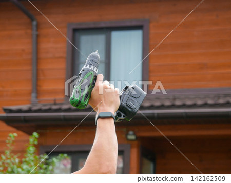 A person's hand raising a green and black cordless drill upward against the backdrop of a wooden house facade. A person's hand raising a green and black cordless drill upward against the backdrop of a wooden house facade. 142162509