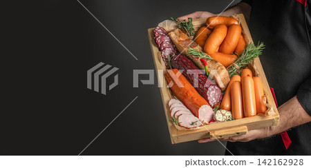 Wooden tray filled with assorted sausages, salami, and bread, held by a person wearing a black apron against a dark background. space for text Wooden tray filled with assorted sausages, salami, and bread, held by a person wearing a black apron against a dark background. space for text 142162928