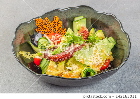 Colorful salad with octopus, fresh vegetables, and decorative elements served in a black bowl on a gray surface Colorful salad with octopus, fresh vegetables, and decorative elements served in a black bowl on a gray surface 142163011