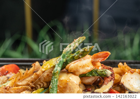 Grilled seafood dish featuring shrimp, octopus, and asparagus garnished with colorful vegetables served on a black plate with a blurred green background Grilled seafood dish featuring shrimp, octopus, and asparagus garnished with colorful vegetables served on a black plate with a blurred green background 142163022