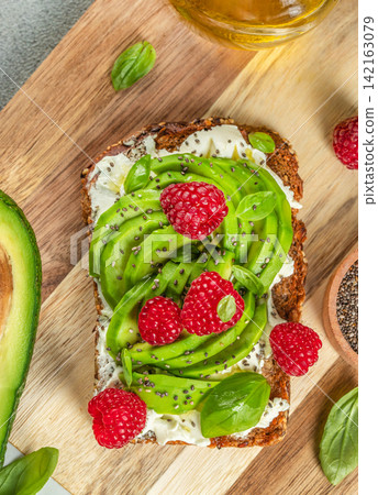 Sliced whole grain bread topped with creamy cheese, avocado, fresh raspberries, and basil leaves on a wooden cutting board with olive oil in the background Sliced whole grain bread topped with creamy cheese, avocado, fresh raspberries, and basil leaves on a wooden cutting board with olive oil in the background 142163079