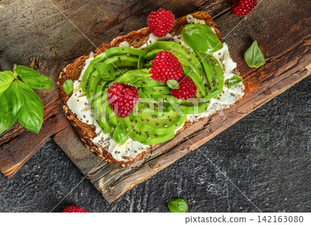 Avocado toast topped with fresh raspberries and basil leaves on a rustic wooden board against a dark textured background Avocado toast topped with fresh raspberries and basil leaves on a rustic wooden board against a dark textured background 142163080