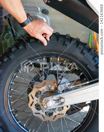 Man mechanic inspecting tread on motorcycle tire in garage. Professional repairman checking rubber wheel condition. Maintenance and service of off road bike for adventure journey. Man mechanic inspecting tread on motorcycle tire in garage. Professional repairman checking rubber wheel condition. Maintenance and service of off road bike for adventure journey. 142163668